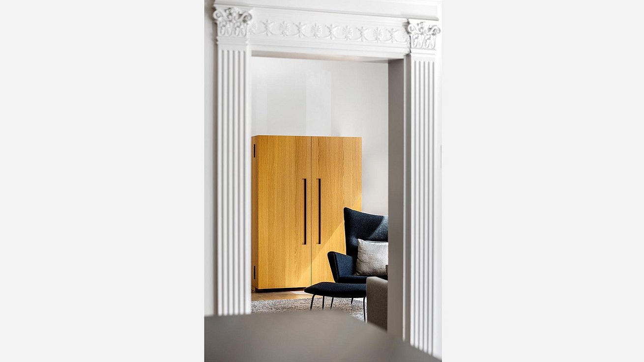 Door frame detail through which you can see a bulthaup b2 kitchen tool cabinet with natural oak finish and a Carl Hansen wing chair and foot rest with dark grey felt fabric.